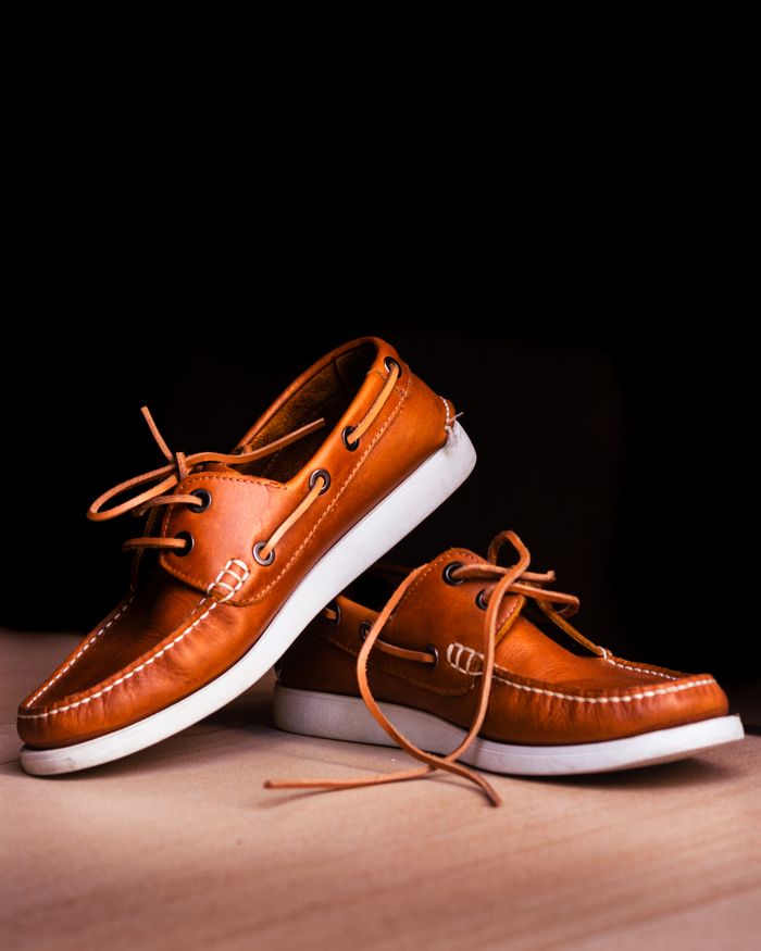 services-01 Close-up of brown leather boat shoes with white soles on a wooden floor against a black background.